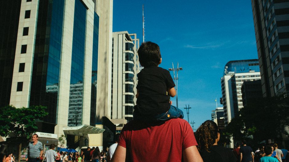 Un padre lleva a su hijo sobre sus hombros a través de una animada calle urbana llena de altos edificios.