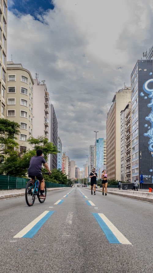 Free A lively street in São Paulo, Brazil, with people cycling and jogging under a clouded blue sky. Stock Photo