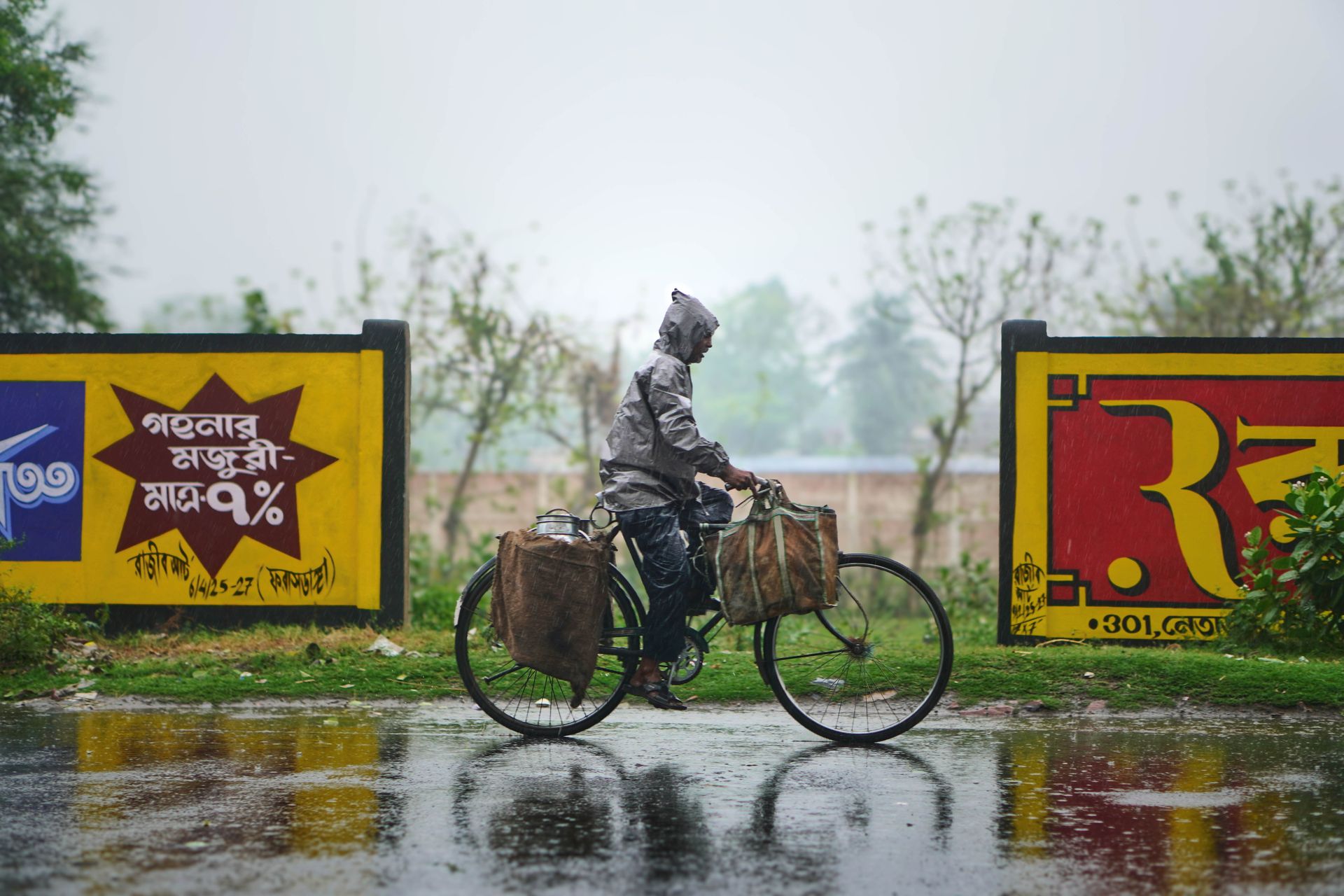 A cyclist rides through the rain by vibrant advertising billboards in a city setting, creating a lively urban scene.