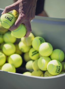 Close-up of a hand grabbing tennis balls from a basket outdoors.