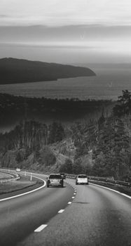 Black and white photo of cars driving along a coastal highway with ocean view.