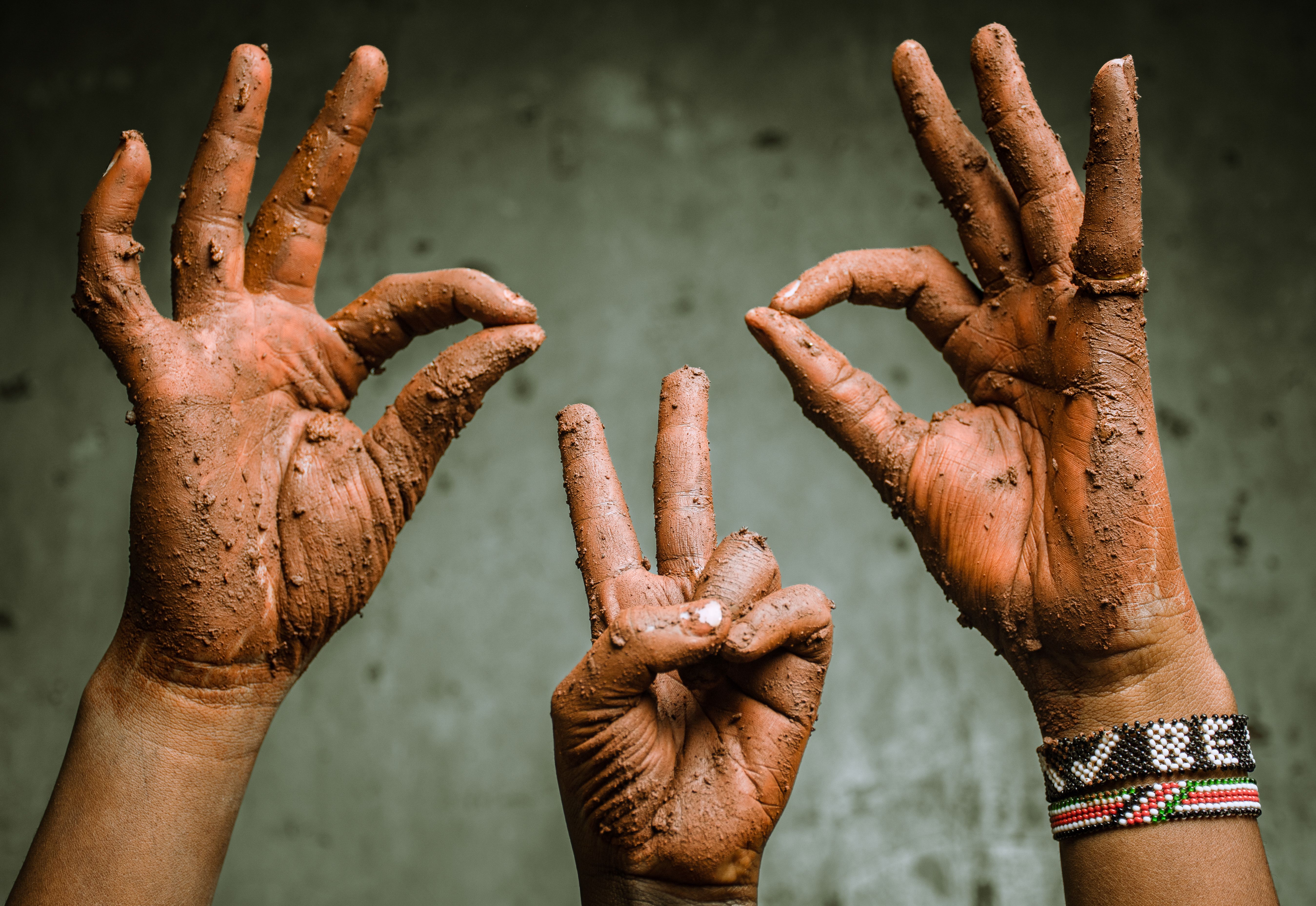 Three clay-covered hands making peace and okay signs outdoors in Nairobi, Kenya.
