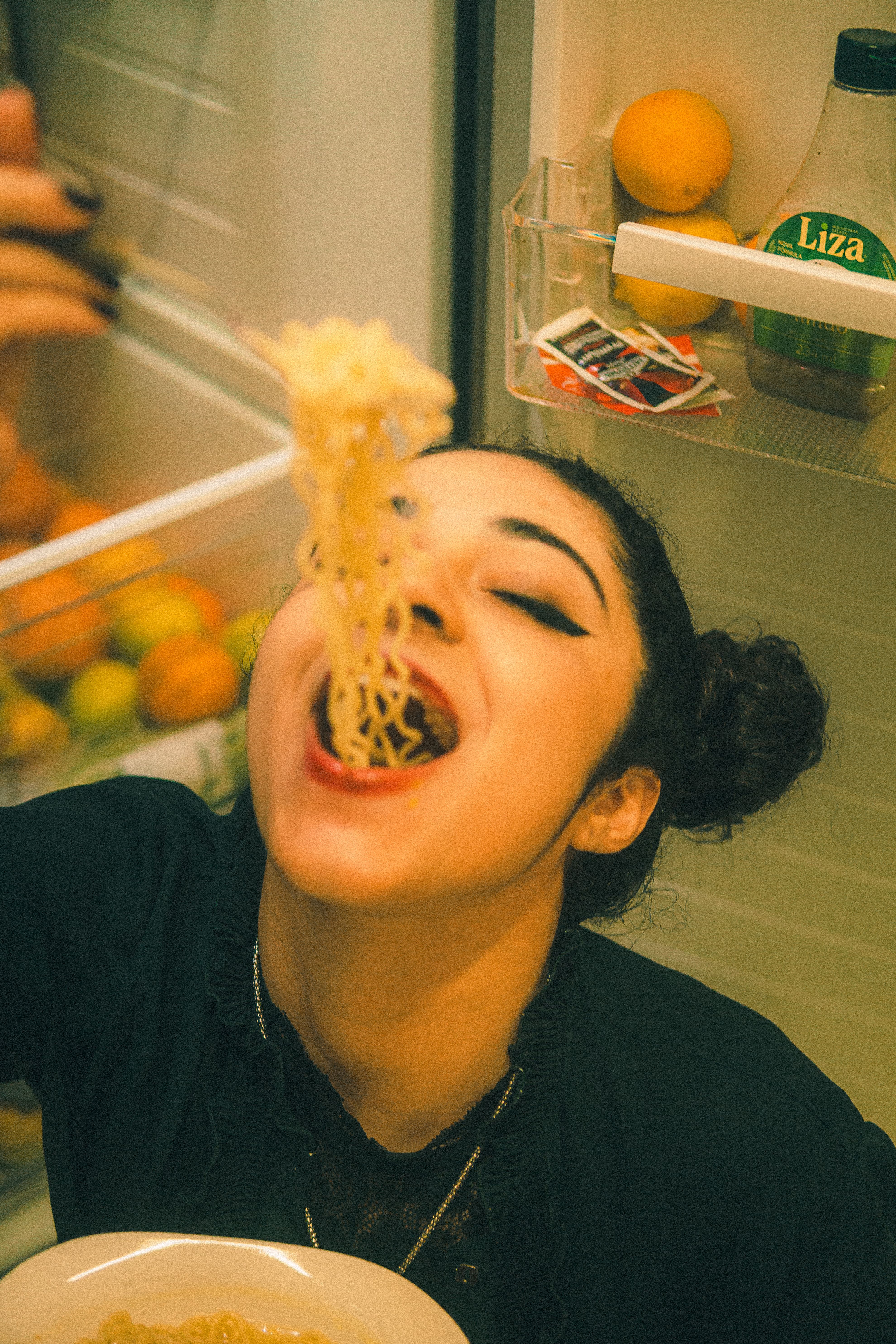 Woman eating instant noodles in front of an open fridge, capturing late-night cravings.