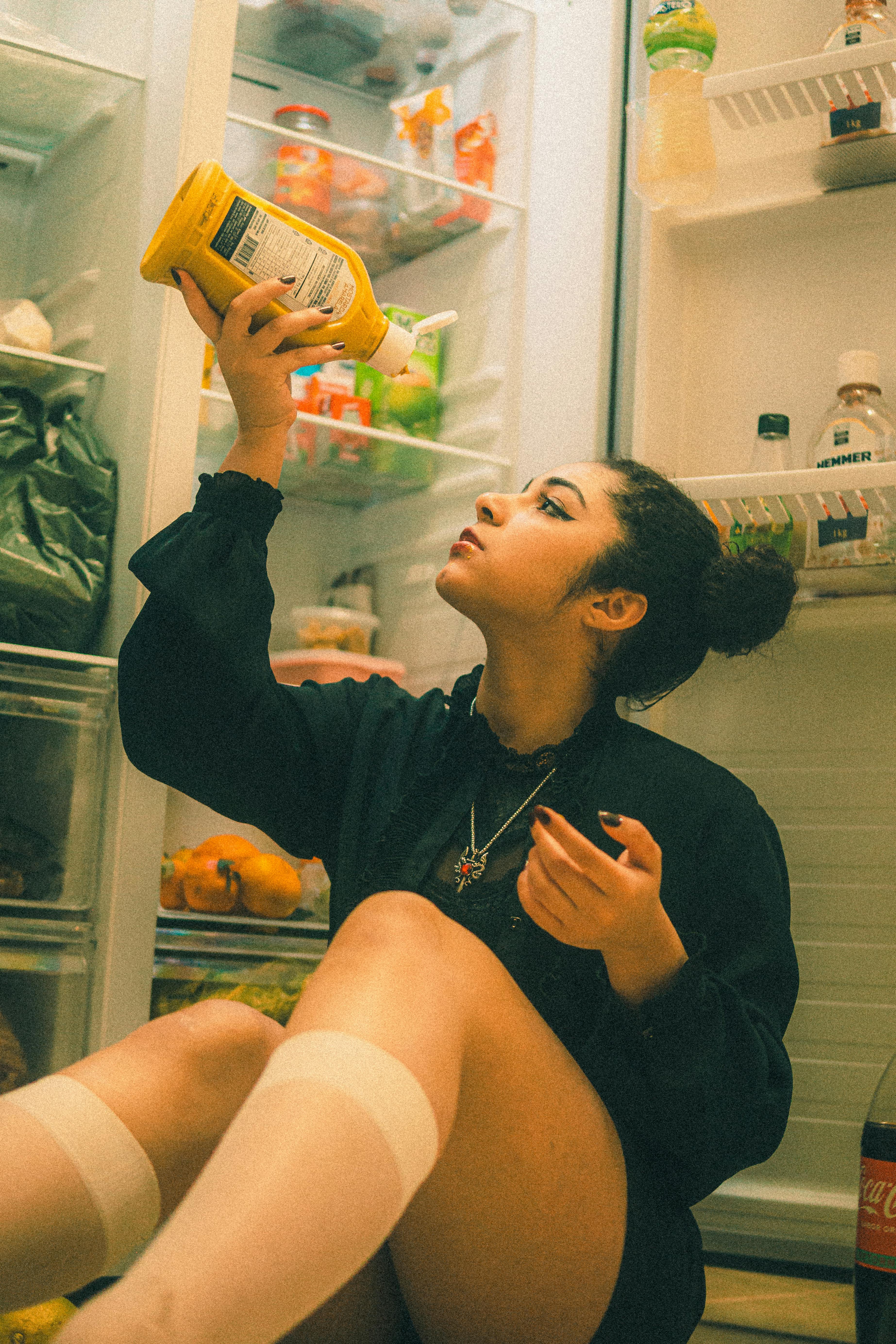 Woman sitting in front of an open fridge at night, examining food jar contents.