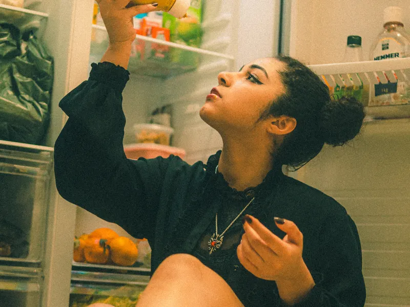 Woman sitting before an open refrigerator at night checking a food jar Woman sitting before an open refrigerator at night checking a food jar