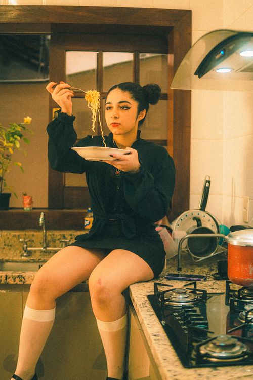 Free A young woman enjoys noodles while sitting on a kitchen counter, creating a cozy indoor scene. Stock Photo