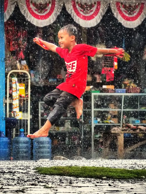 Free A happy child in a red shirt jumps with joy in the rain on a street in West Java, Indonesia. Stock Photo