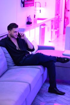 Stylish man in black outfit sitting on a sofa with pink neon lighting backdrop.