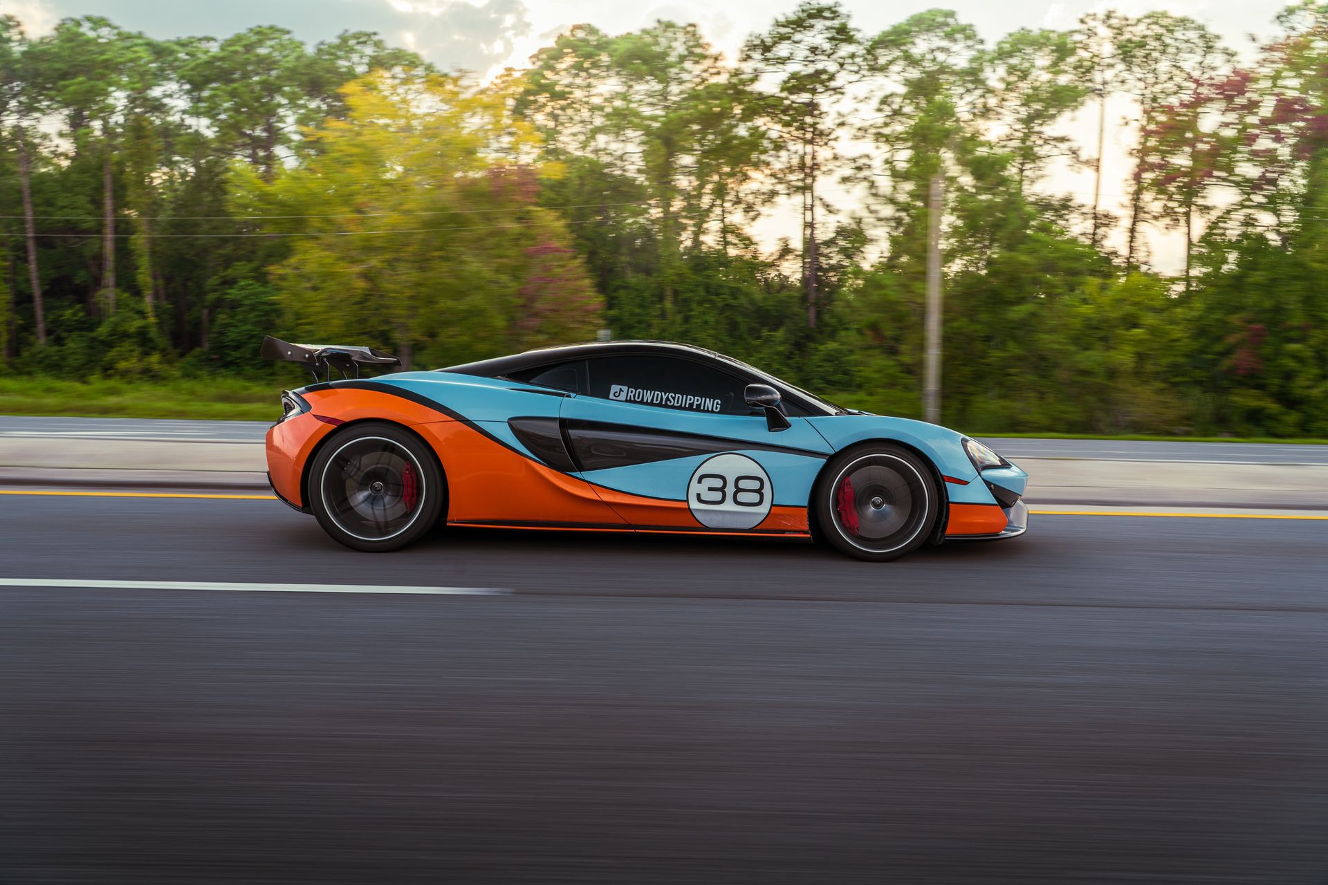 Dynamic shot of a sports car with Gulf livery racing down a tree-lined highway.