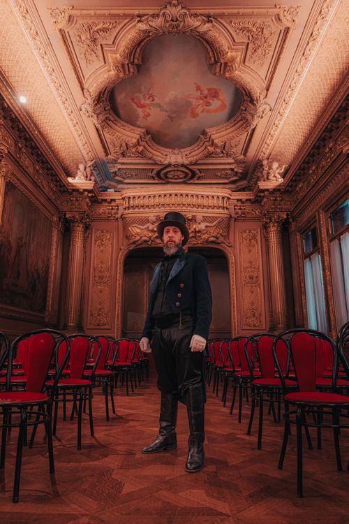 Free A man in vintage attire stands in an ornate room with red chairs and a painted ceiling. Stock Photo