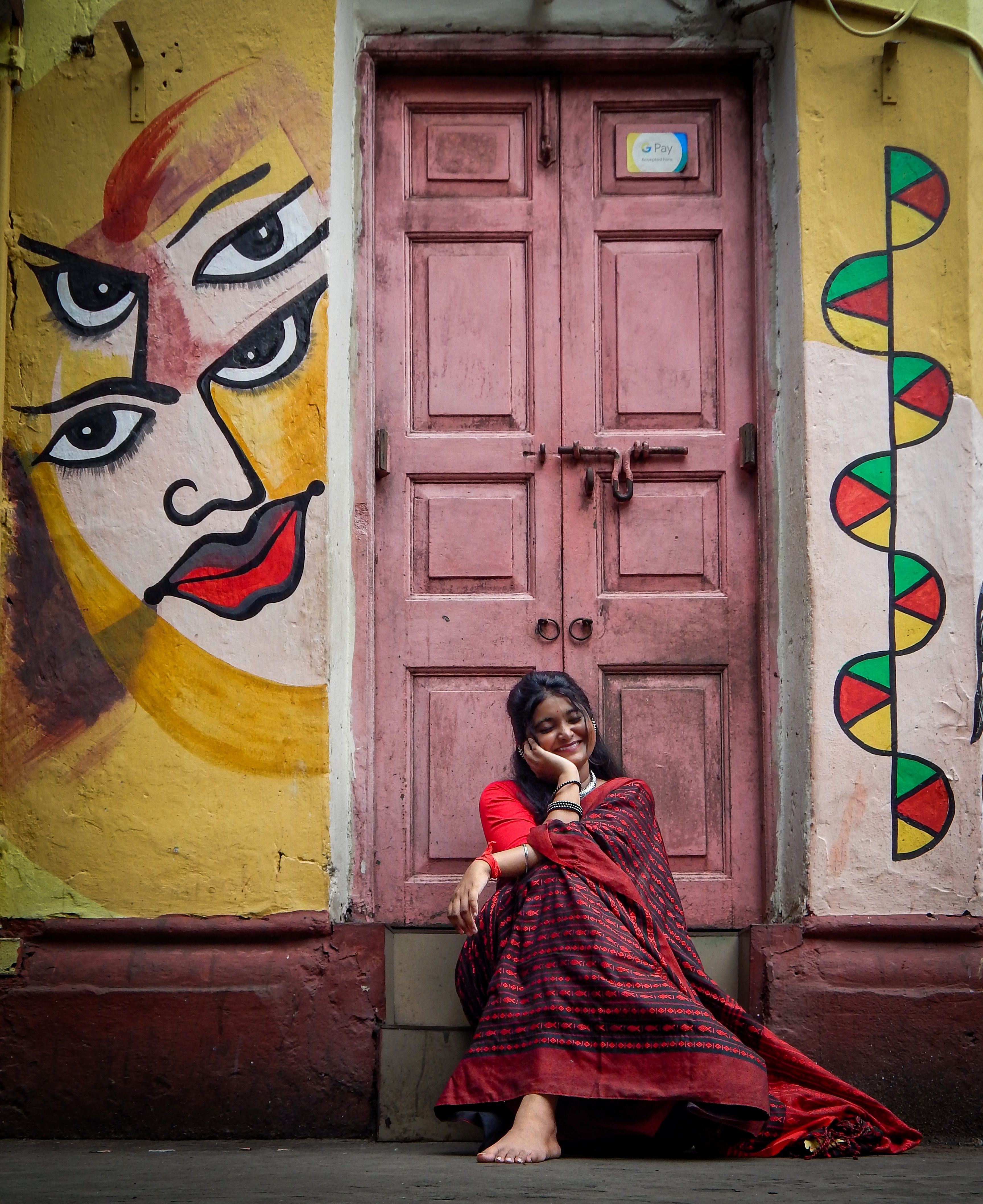 Smiling woman in red saree sits by a colorful, art-adorned doorway in India, embodying cultural charm.