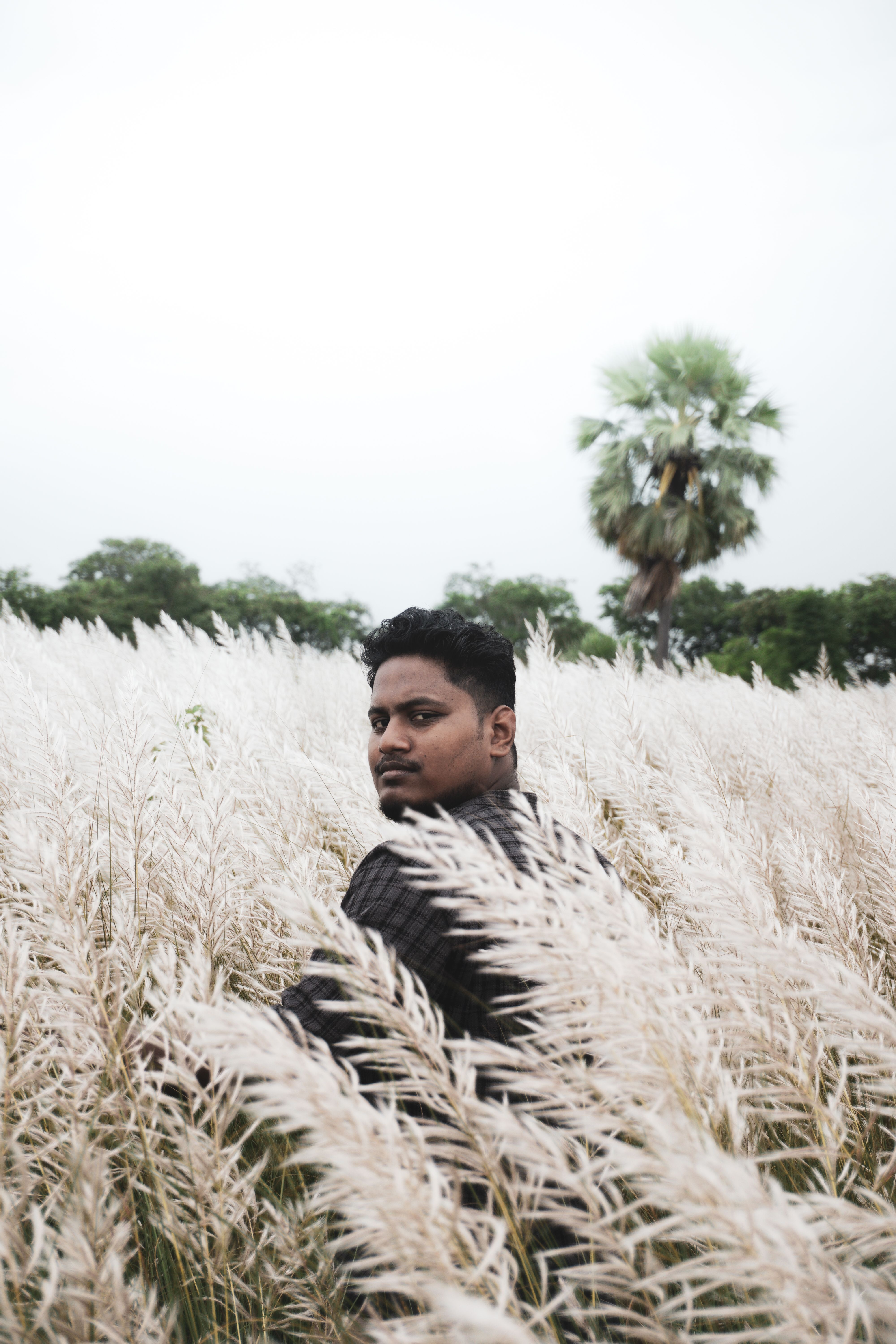 A man standing in a field of tall grass, glancing back toward the camera under a cloudy sky.