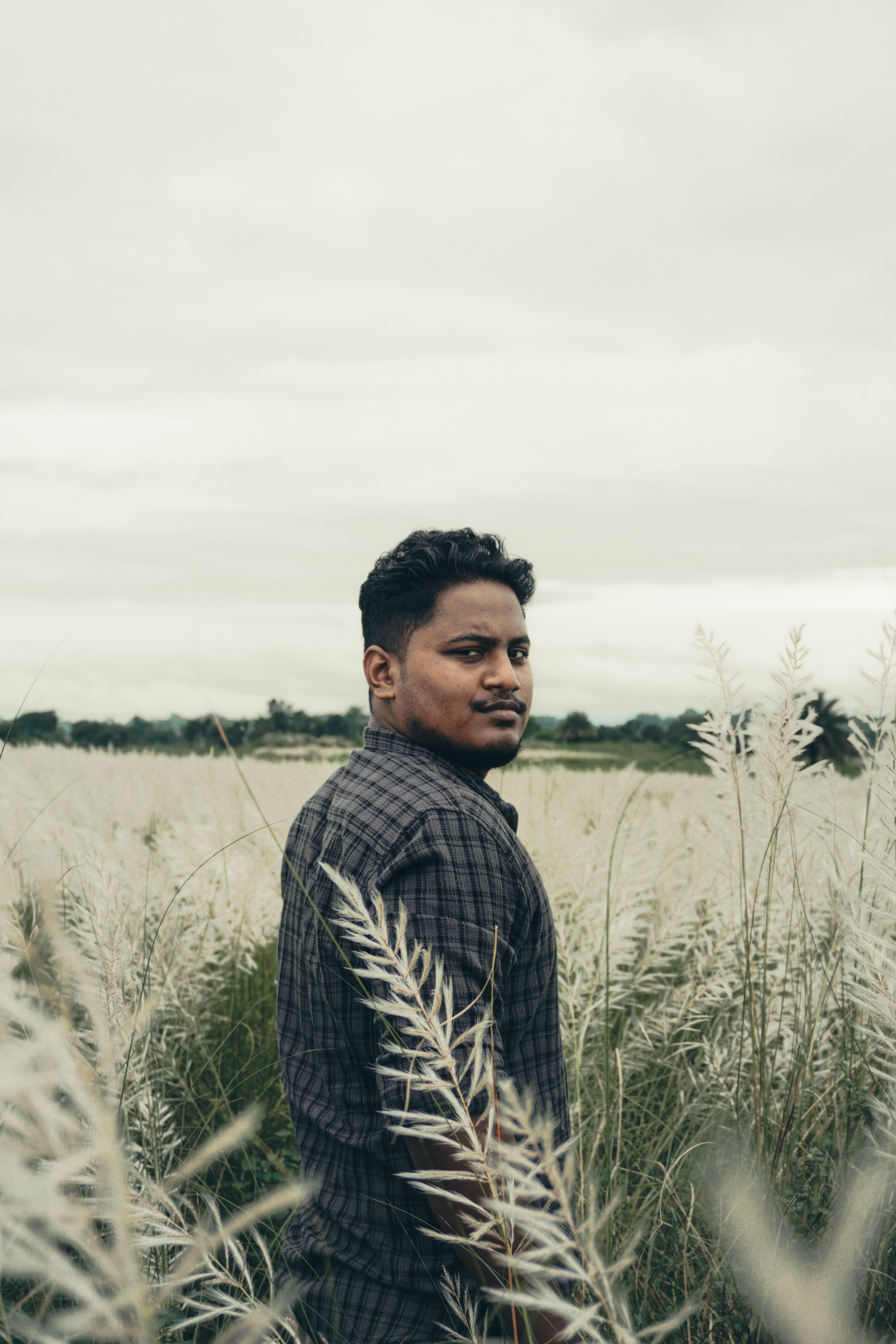 A man in a plaid shirt standing in a vast field with tall green grasses under a cloudy sky.