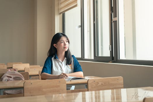 Young student in uniform sitting at a classroom desk, looking thoughtful by the window.
