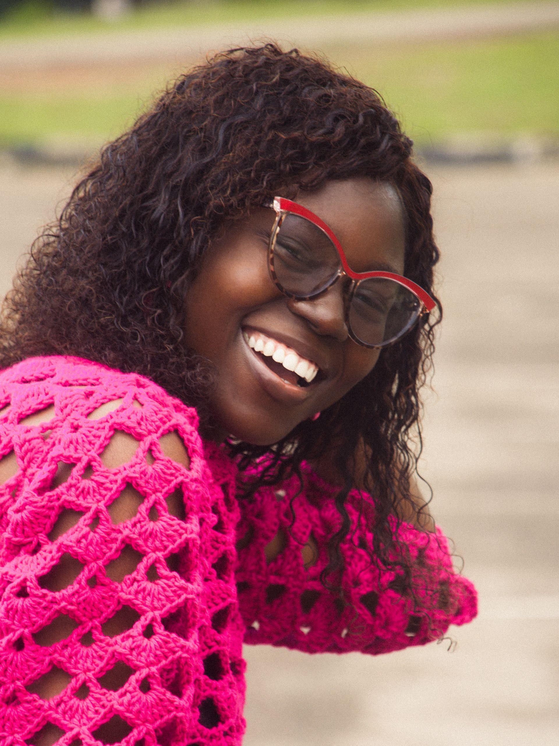 Cheerful woman with curly hair wearing glasses and a vibrant pink sweater, smiling outdoors.