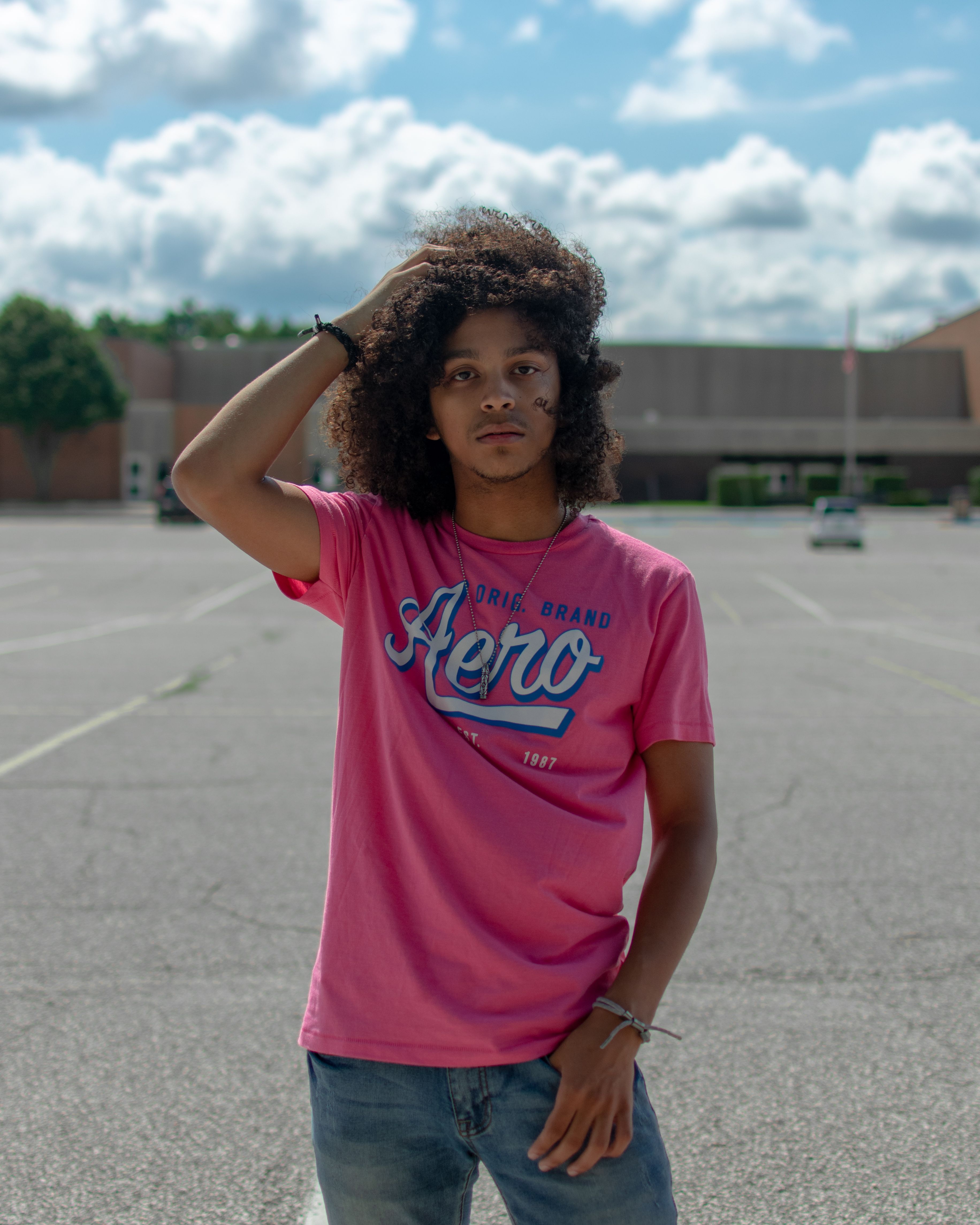 Young man posing in a parking lot wearing a pink t-shirt on a sunny day.