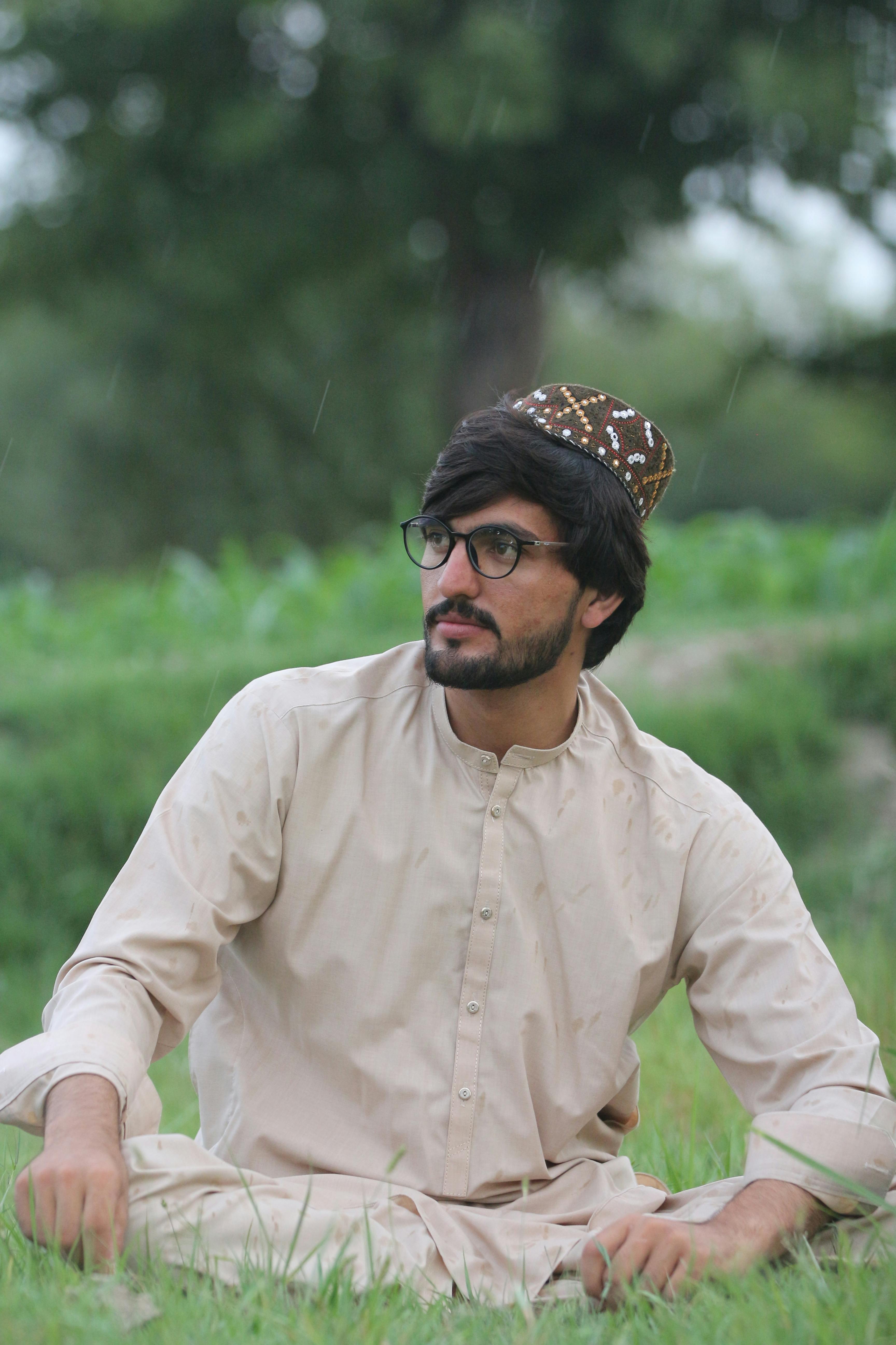 A young man in traditional attire seated outdoors on a grassy field with a thoughtful expression.