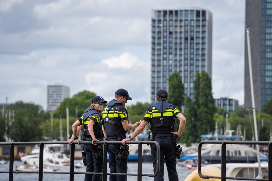 Three police officers stand on a waterfront railing, overseeing a cityscape with high-rise buildings.