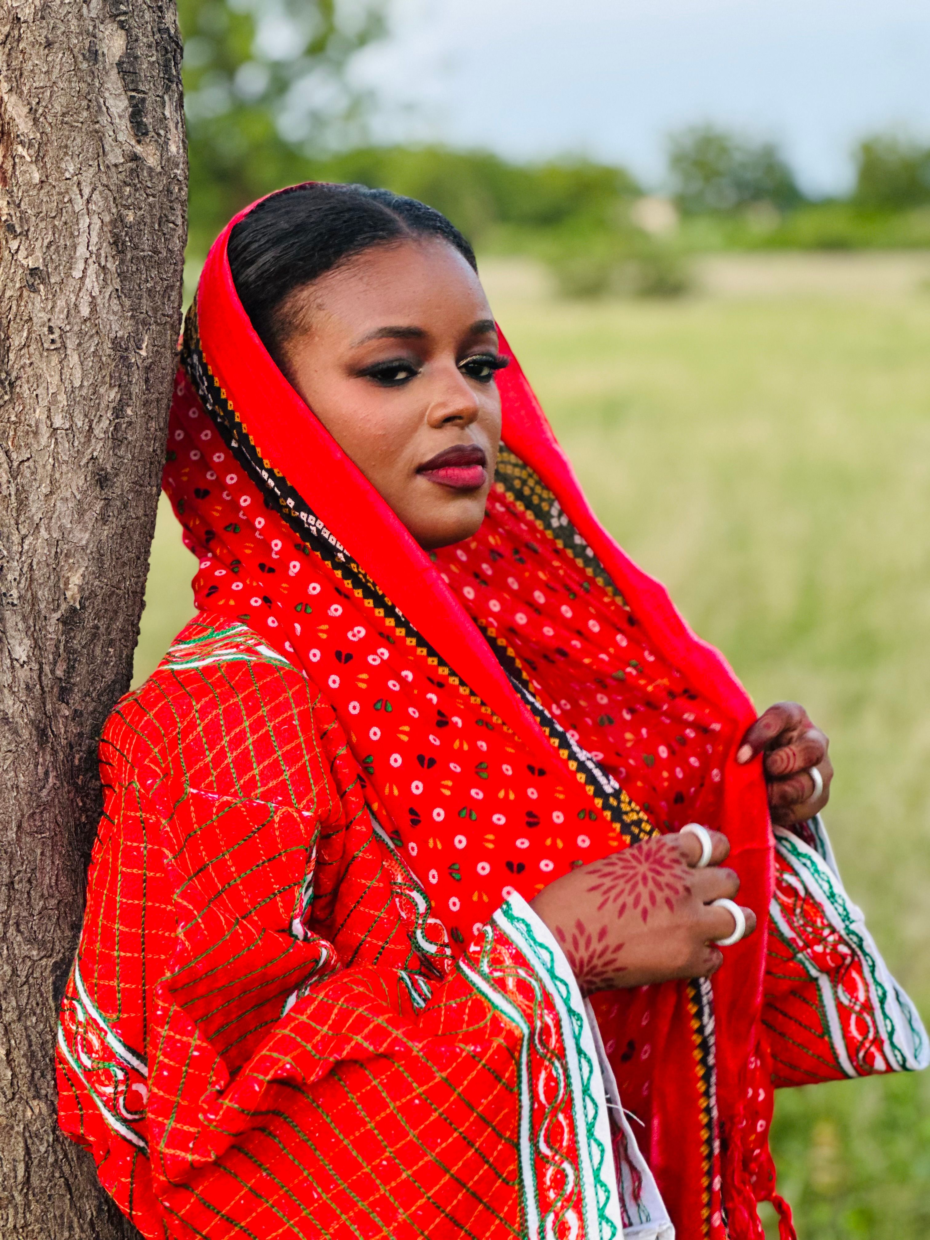 Woman in vibrant red traditional clothing leaning against a tree in a serene outdoor setting.
