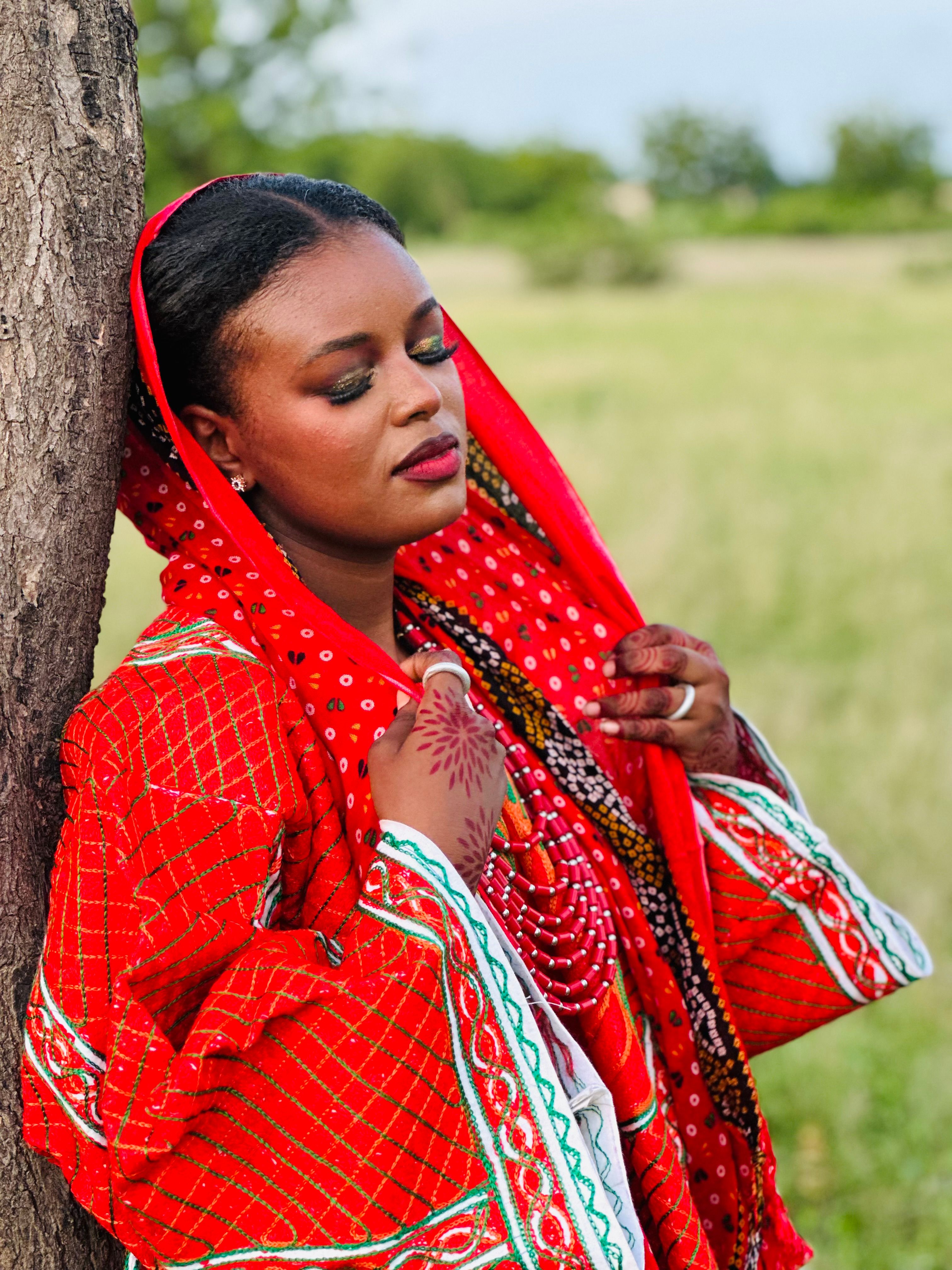 Woman in vibrant red traditional clothing with henna leaning on a tree.