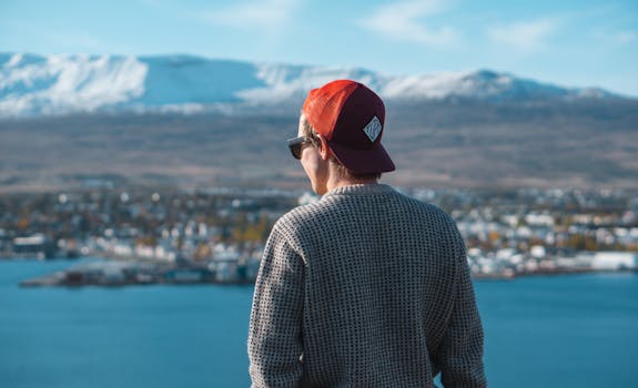 A man wearing a red cap and sunglasses observes a scenic city view with snow-covered mountains.
