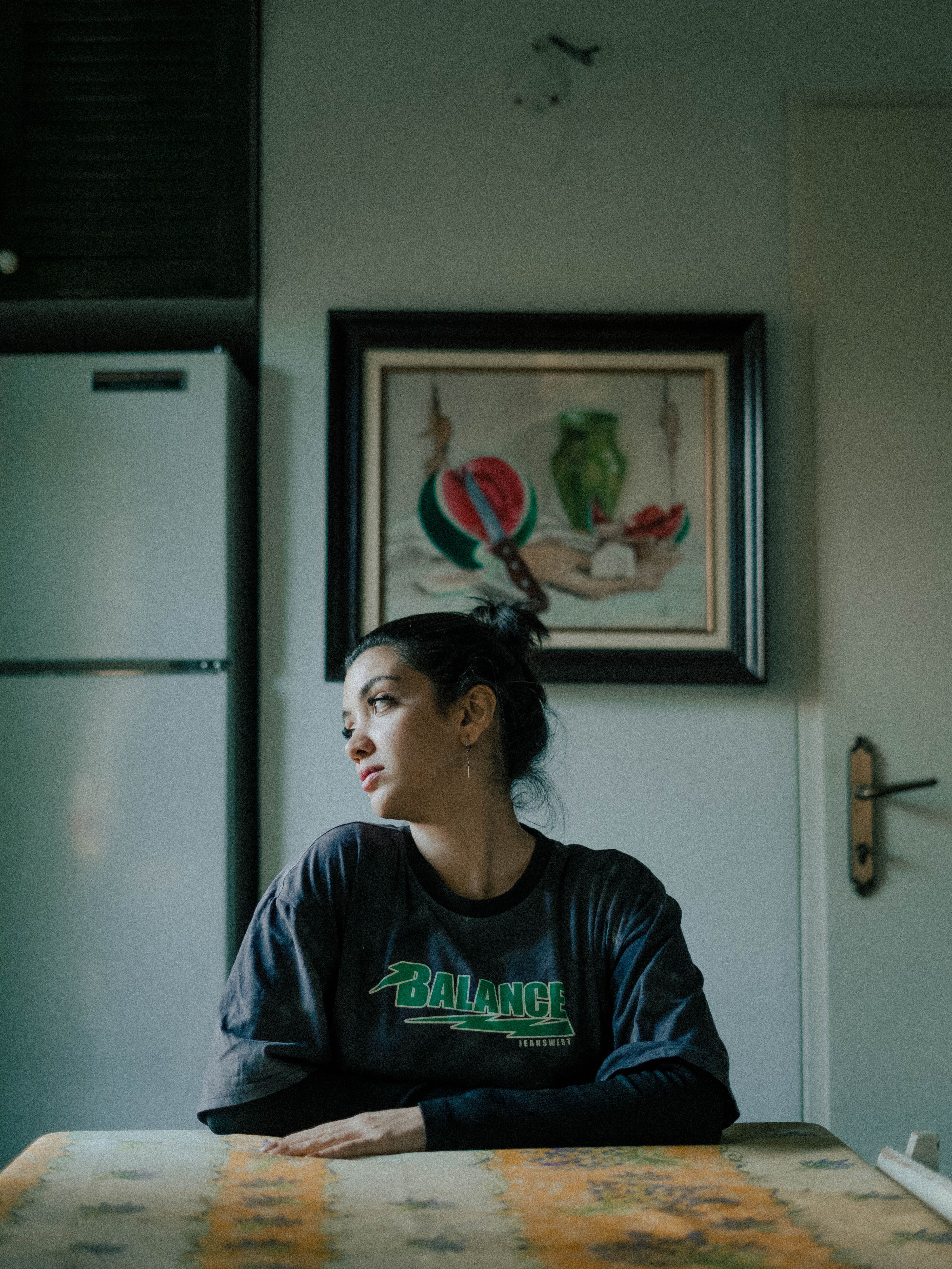 A woman sitting at a kitchen table, looking contemplative with a painting on the wall behind.