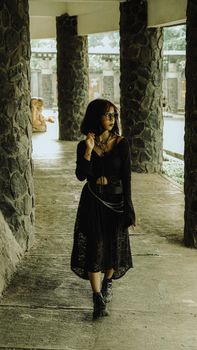 Woman in a black gothic outfit walking under stone columns in an outdoor setting.