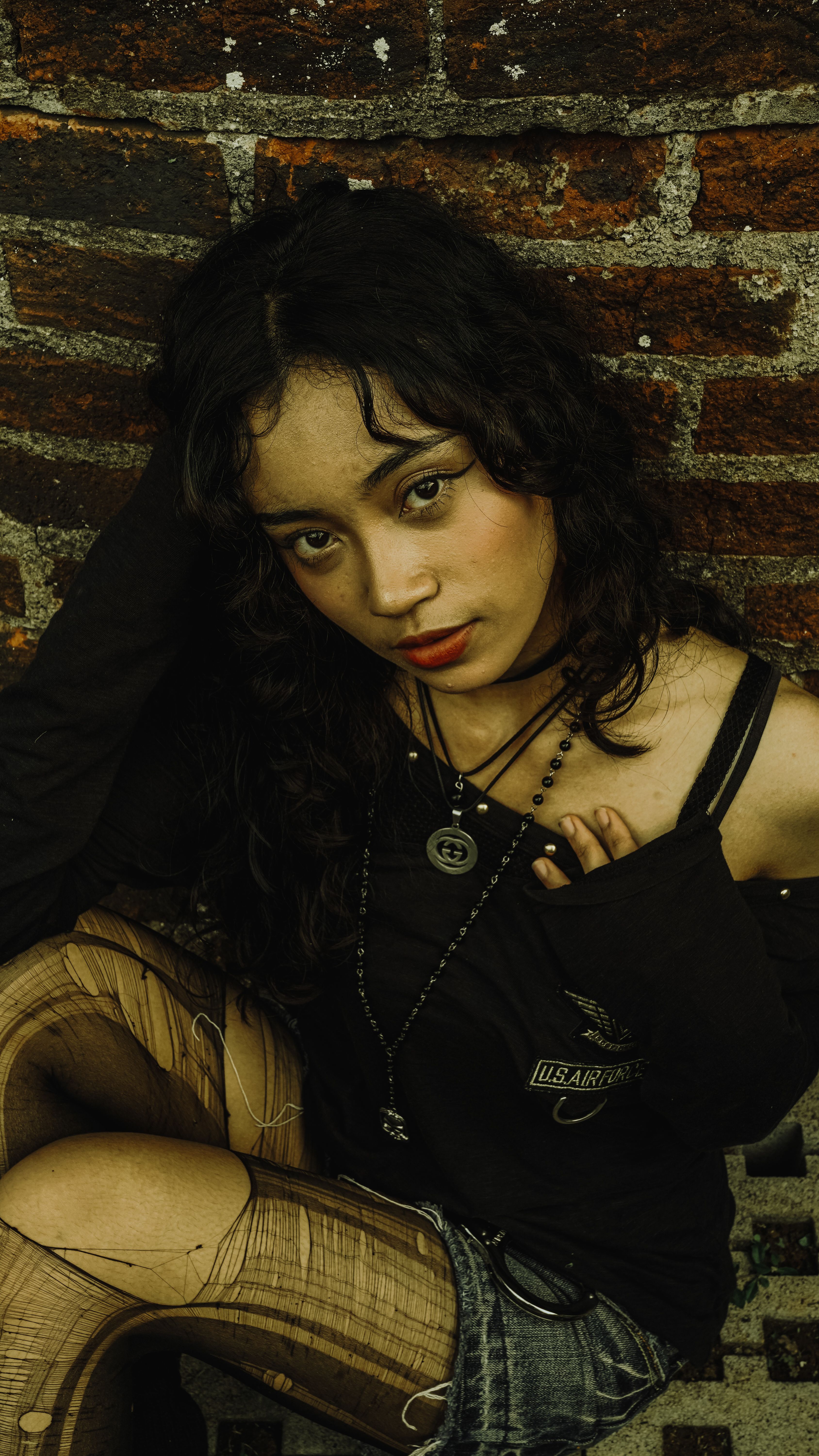 Stylish young woman with an edgy look leans against a brick wall posing confidently.