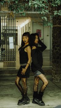 A portrait of two fashionable women in urban attire, standing back to back indoors with greenery overhead.