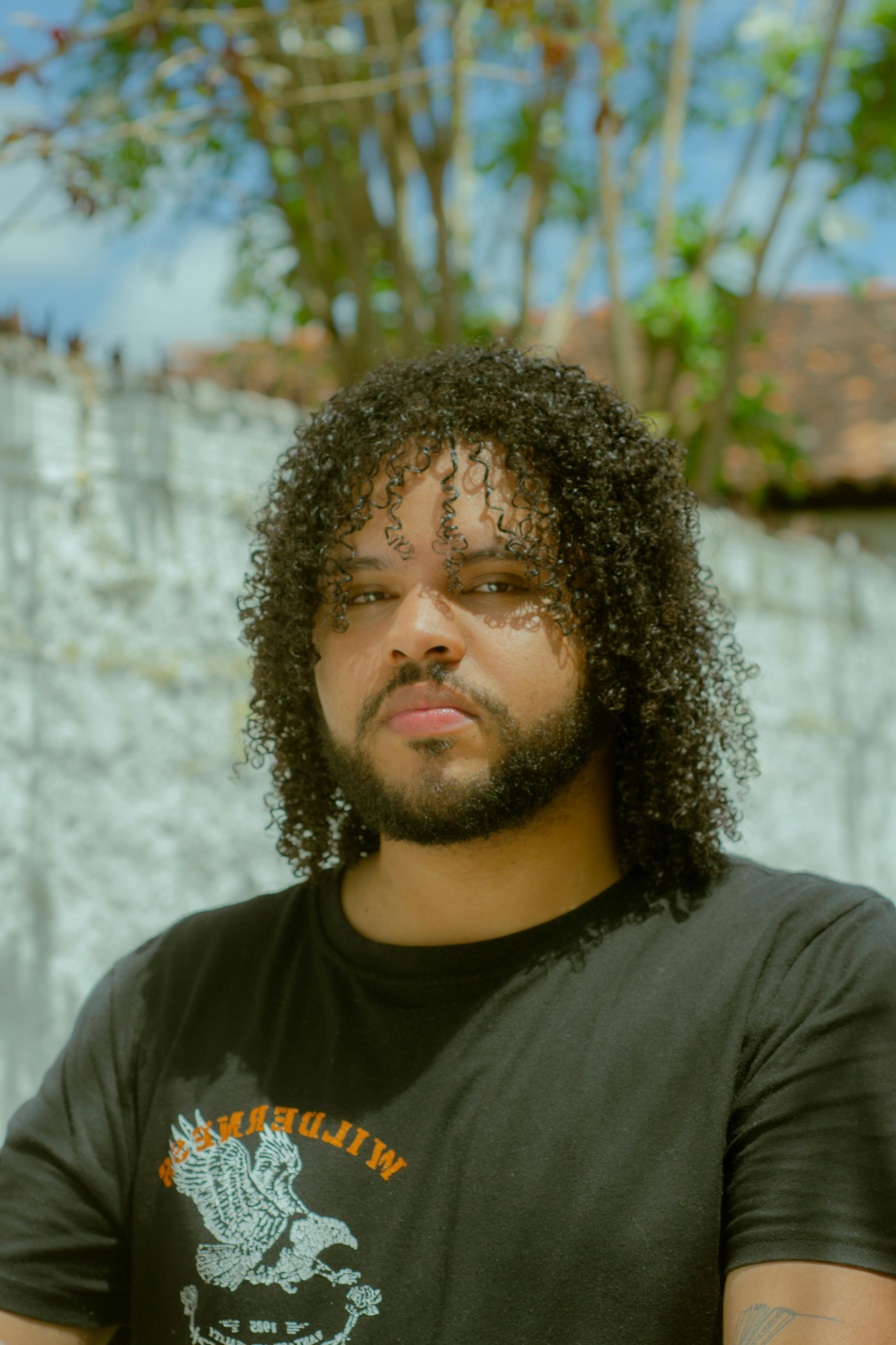 Bearded man with curly hair in a t-shirt poses outdoors against a sunny backdrop.