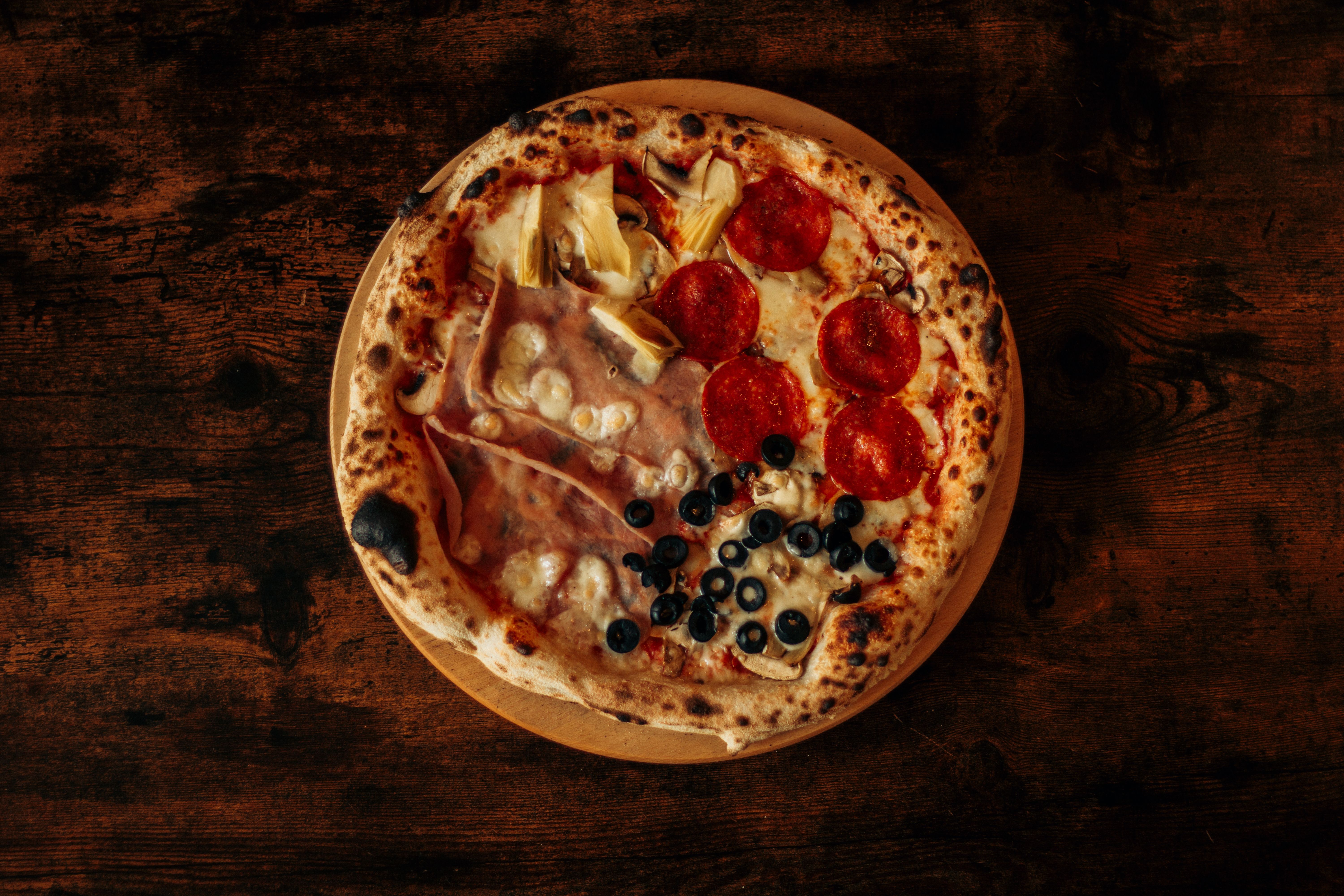 Ilmainen Herkullinen gourmet-pizza, jossa on erilaisia täytteitä, kuten pepperonia, oliiveja ja artisokkaa, maalaismaisella puupöydällä. Kuvapankkikuva