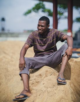 Casual portrait of a smiling man sitting on sand wearing relaxed clothing outdoors.
