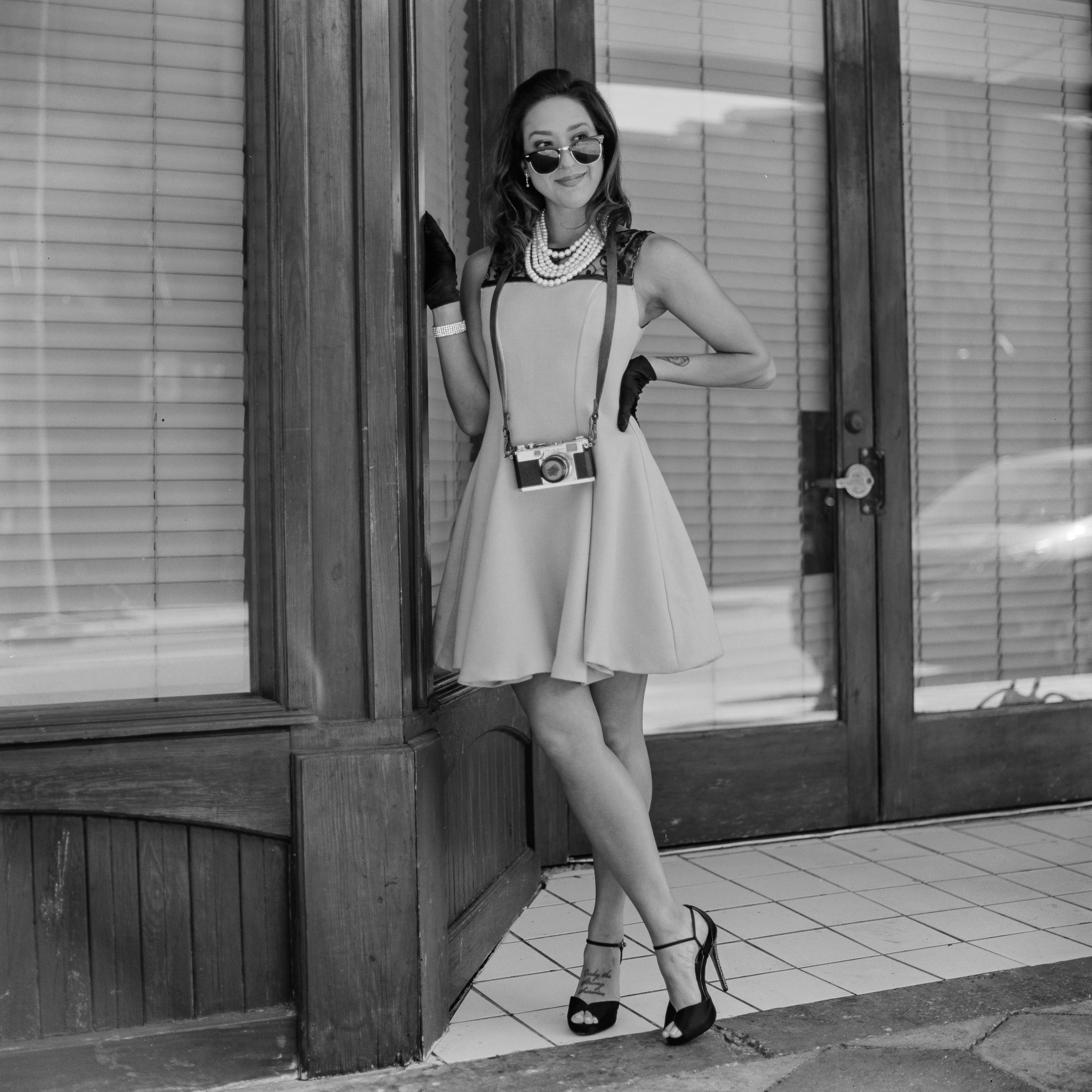 Elegant woman in vintage attire posing with camera outside storefront.