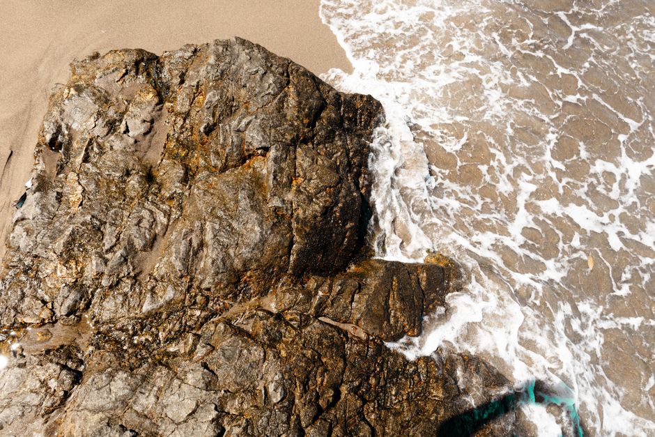 Aerial view of waves crashing against rocky shoreline on a sunny day.