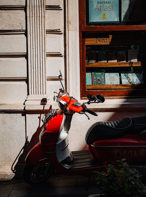 Red vintage scooter parked outside a bookstore with a sunlit facade, conveying a nostalgic urban scene.