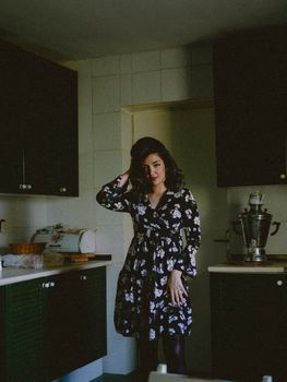 A woman in a floral dress poses in a dimly lit vintage kitchen, creating a moody atmosphere.