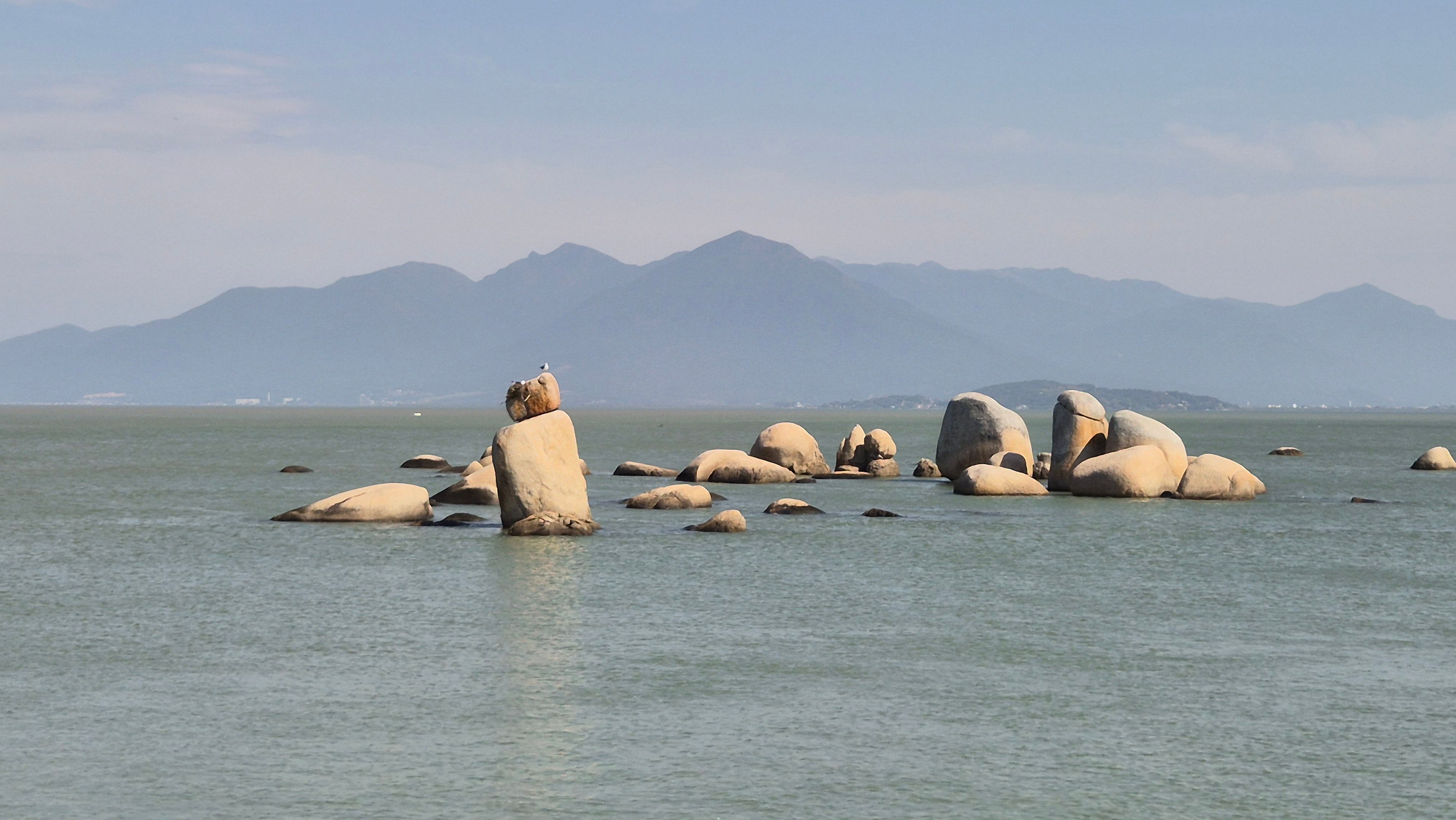 Serene view of large boulders in calm sea with distant mountains under a clear sky.