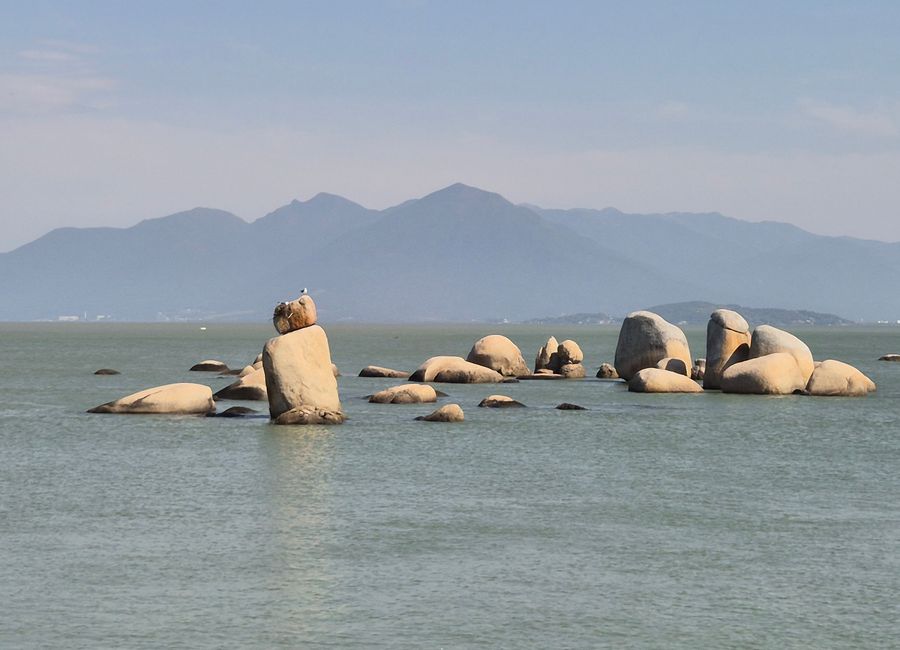 Serene view of large boulders in calm sea with distant mountains under a clear sky.