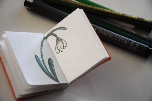 A detailed sketch of a snowdrop flower in a miniature sketchbook with artistic drawing tools in the background.