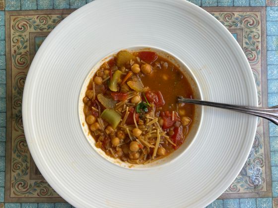 Tomato broth with chickpeas, broken vermicelli, celery and red pepper in a wide white bowl