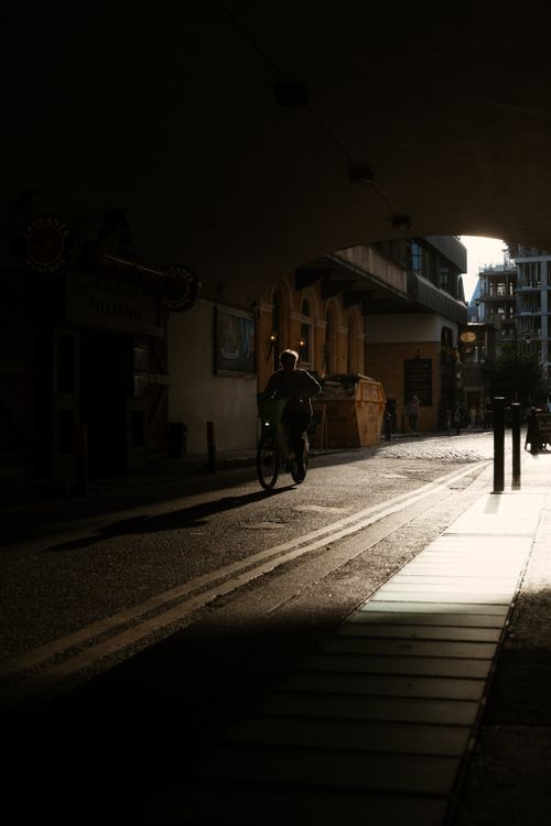 Free A cyclist pedals through a sunlit tunnel in London, casting dramatic shadows on the pavement. Stock Photo
