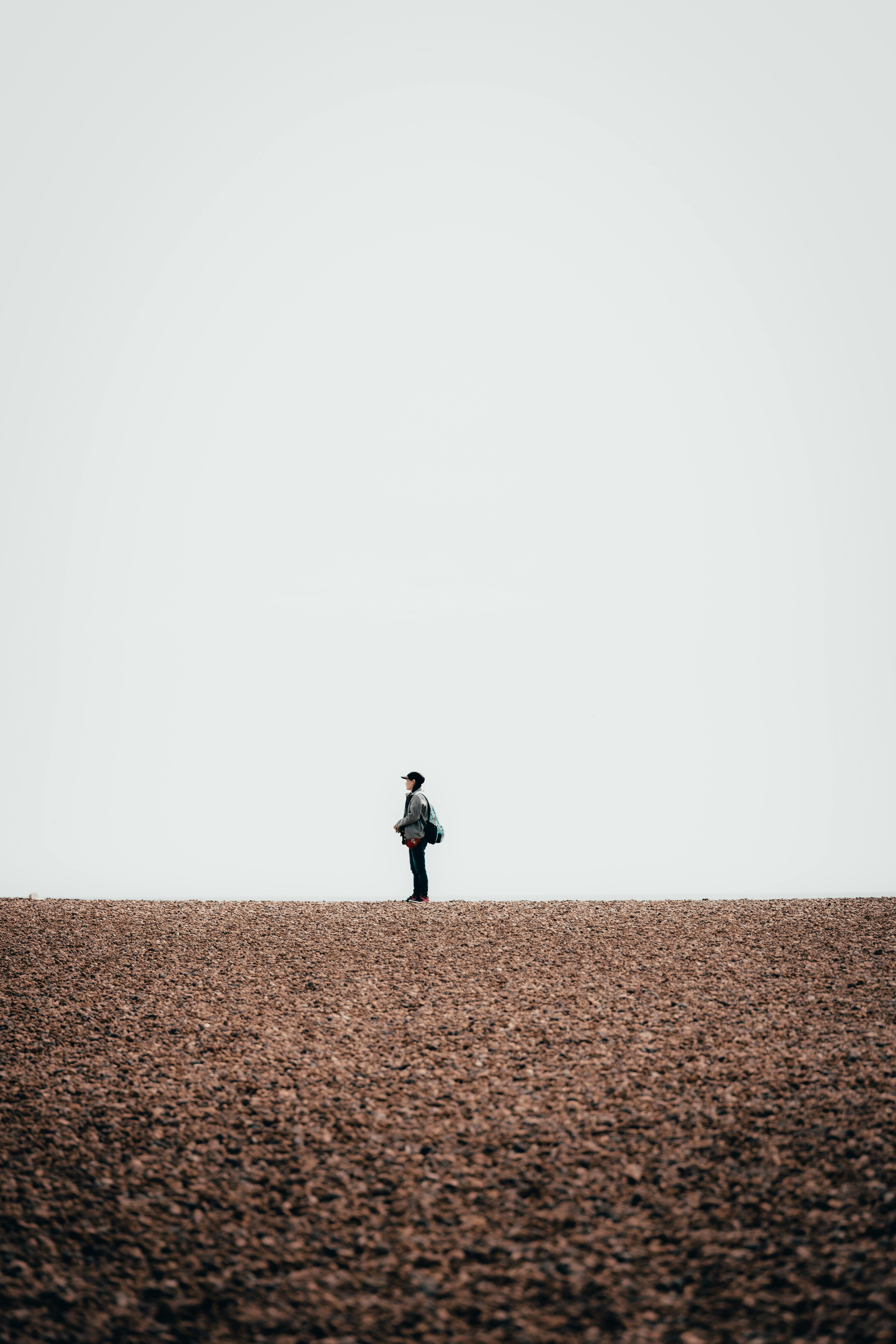 A solitary figure stands on a wide pebble beach at Hove, England, under an expansive sky.