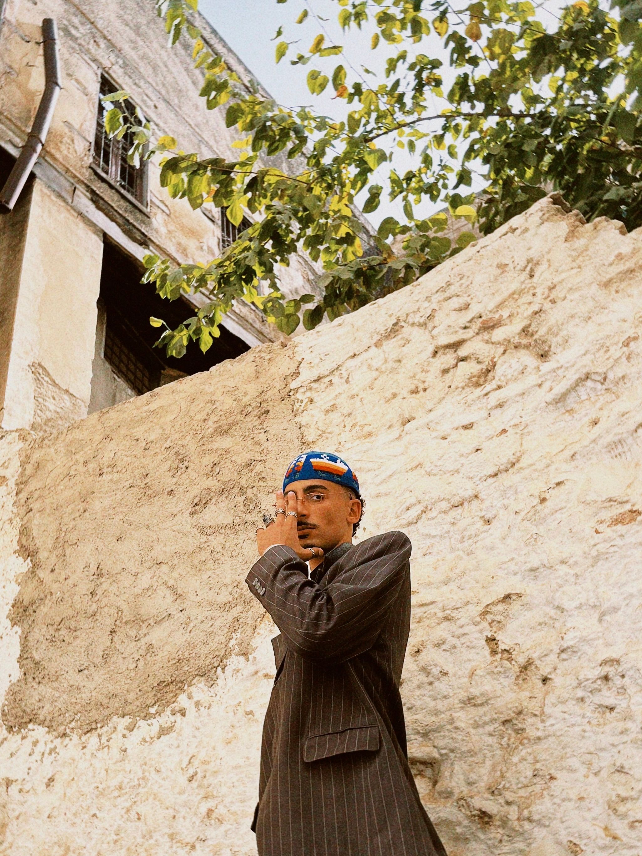 Man in a suit poses by a textured stone wall in urban outdoors.