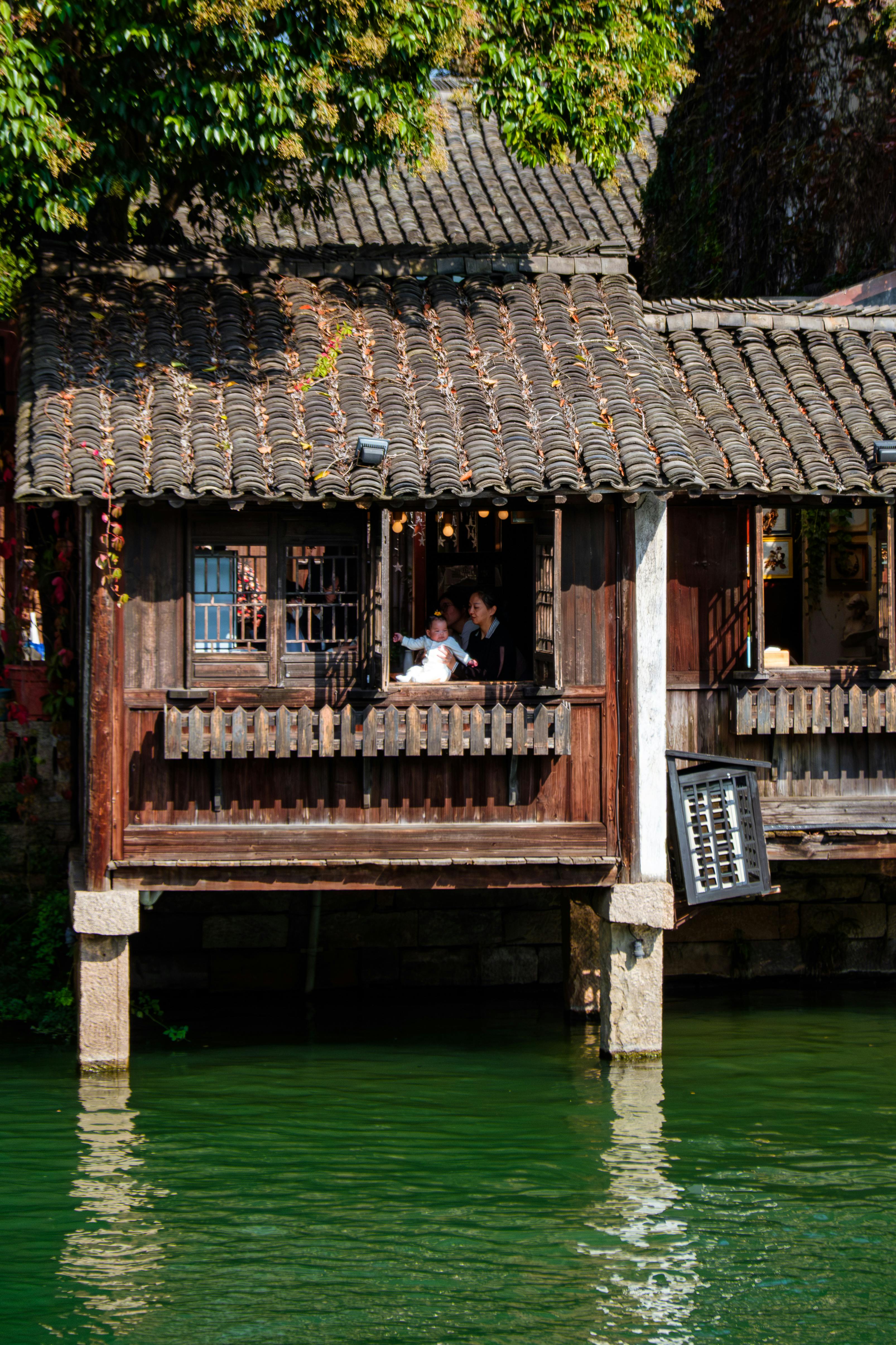 A picturesque Asian wooden house by the river with people on the balcony, reflecting cultural heritage.