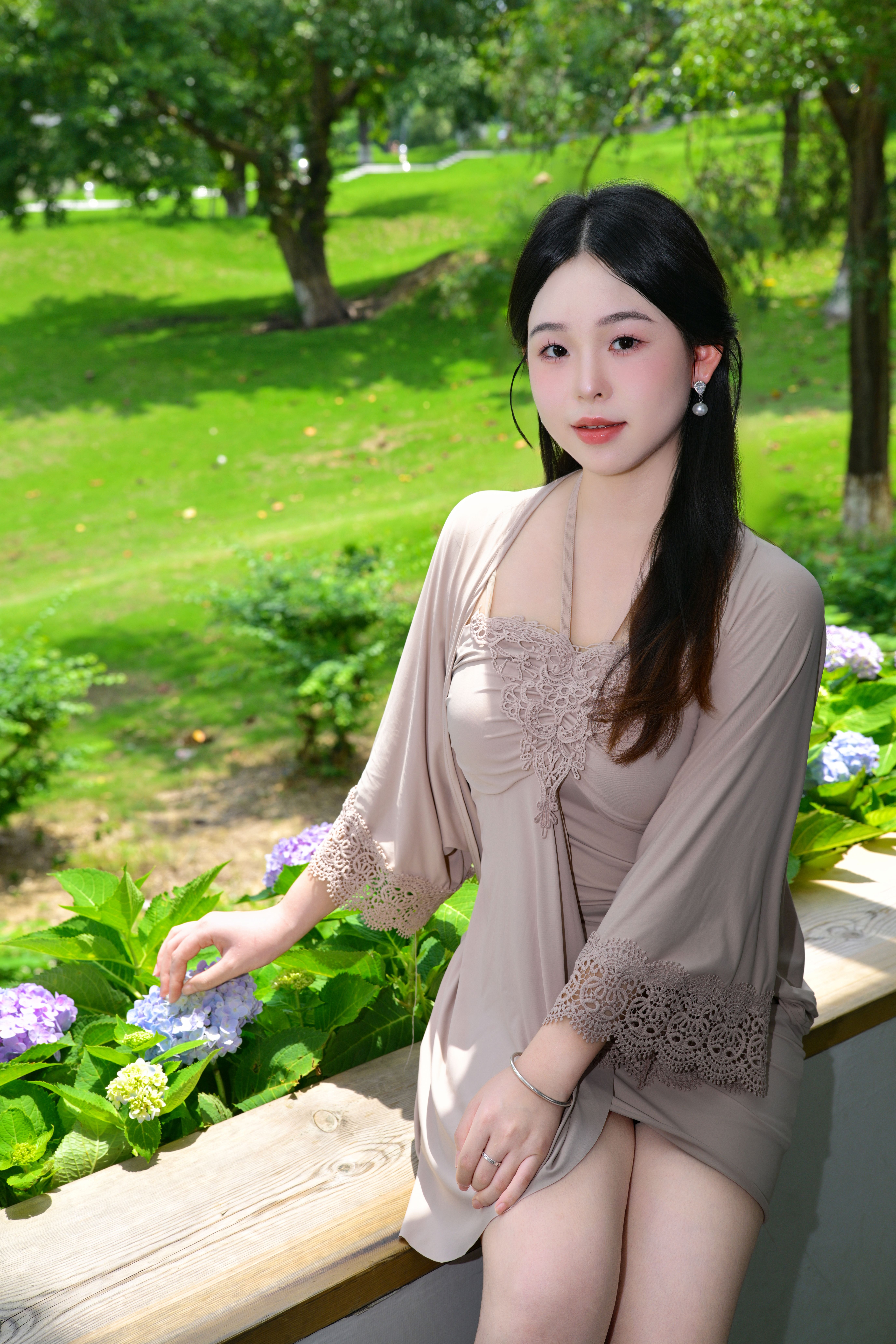 A young woman sitting on a ledge surrounded by greenery and hydrangeas on a sunny day.