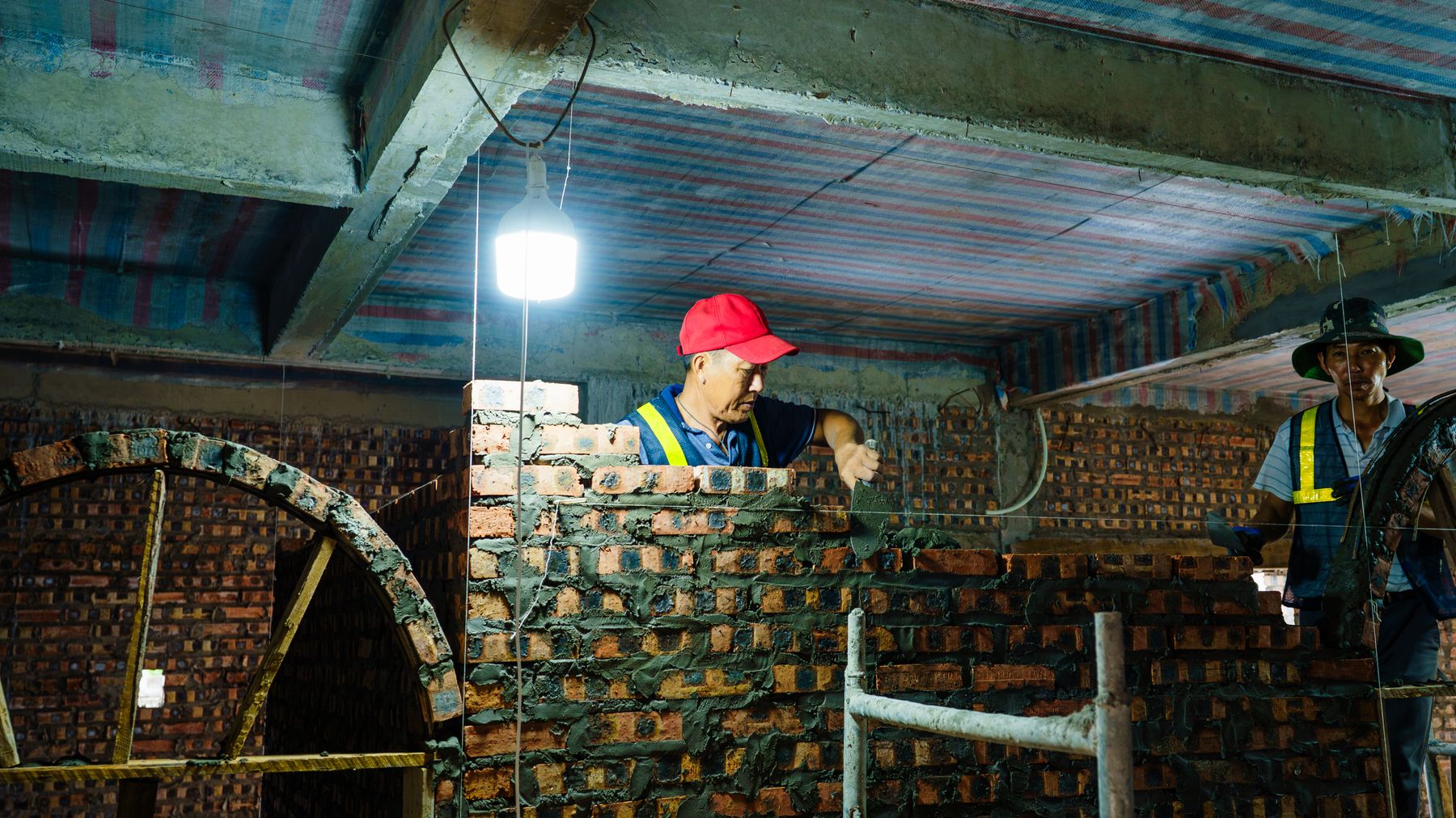 Workers constructing a brick wall indoors under a dimly lit environment in HαΊ£i PhΓ²ng, Viα»t Nam.