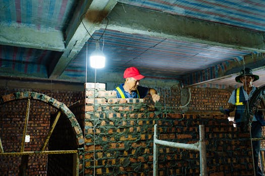 Workers constructing a brick wall indoors under a dimly lit environment in Hải Phòng, Việt Nam.