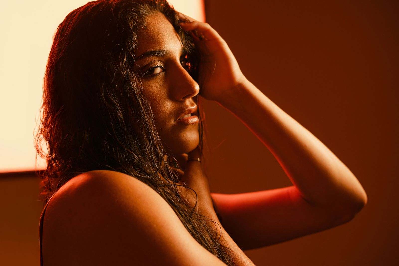 Woman with long wet dark hair, hand raised to her head, lit by warm orange studio light