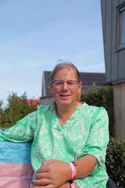 A person in a green top smiling outdoors, holding a trans flag. Bright and positive mood.