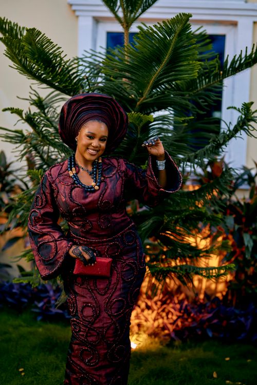 Free Beautiful woman smiling in a maroon traditional African outfit, posing outdoors with vibrant greenery. Stock Photo